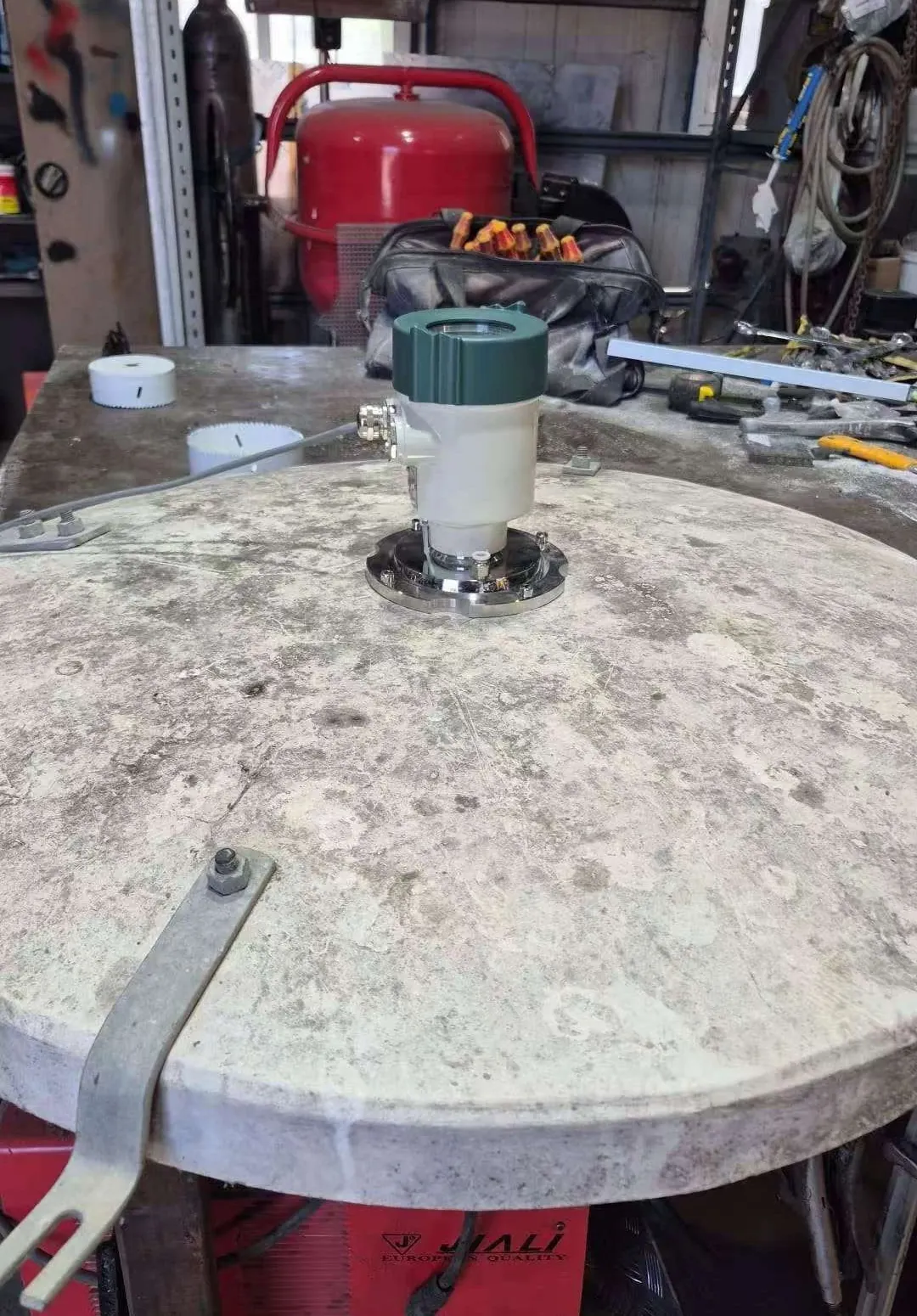 Sensor de nivel de radar de 80 GHz atornillado a través de una brida de acero inoxidable en el centro de una tapa circular de hormigón de silo, montado sobre un banco de trabajo