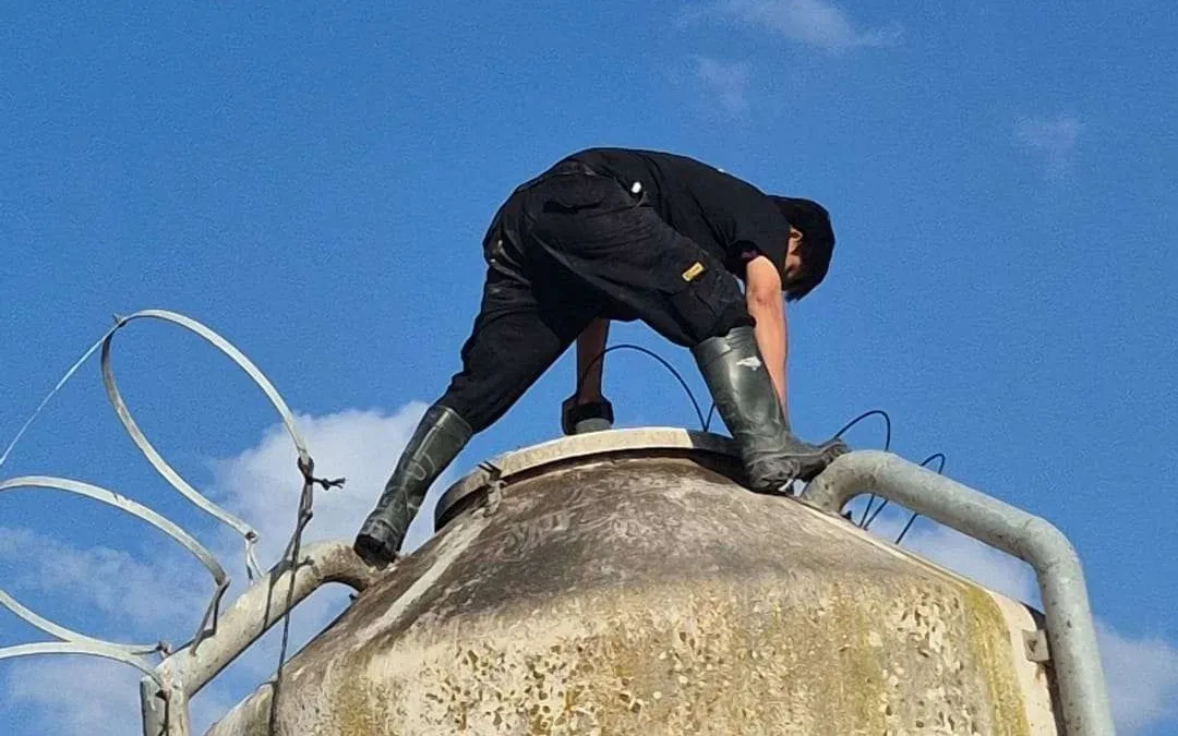 A technician working on the open hatch at the top of an outdoor feed silo, fitting a radar level sensor, against a clear blue sky