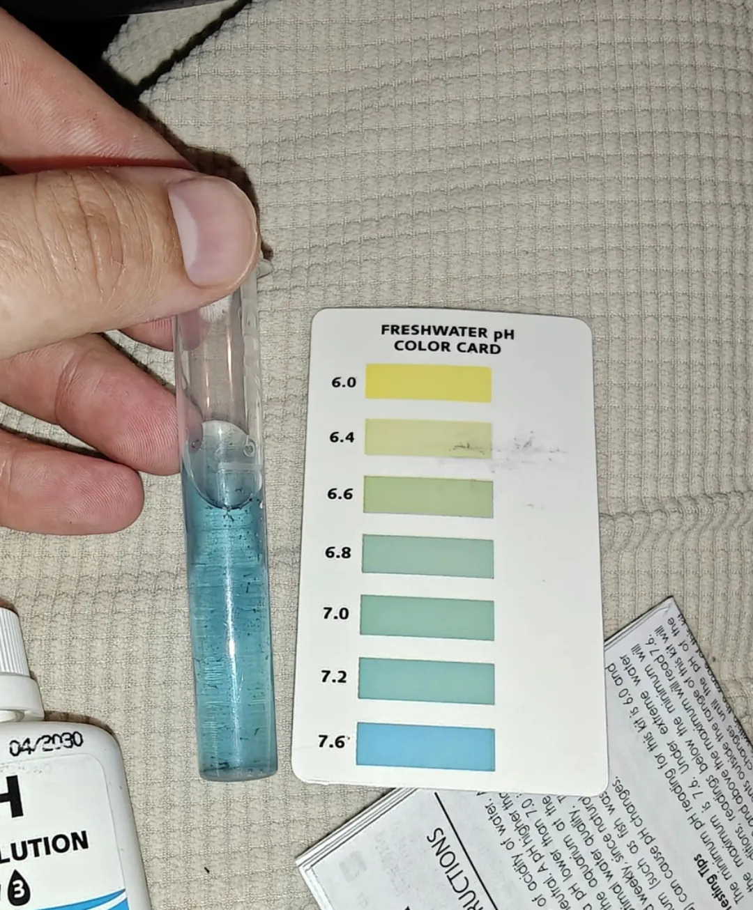 A hand holding a glass test tube of reagent-treated sample next to a printed pH colour comparison card